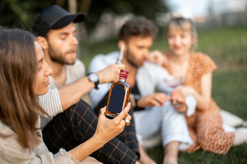 Young friends sit together and hug, talk and drink alcohol in a close and friendly atmosphere on a picnic in the evening. Woman holding bottle with blank label to copy paste