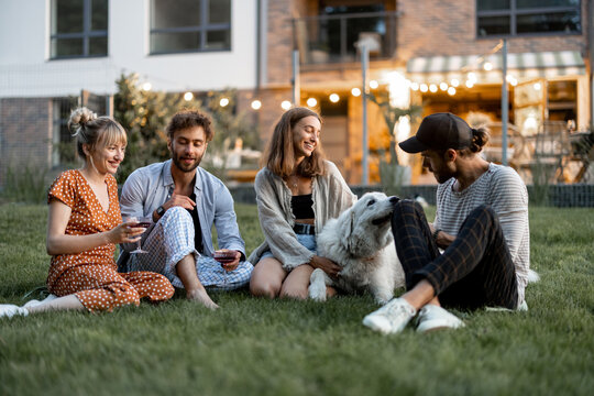 Young friends having fun playing with a dog and drinking wine, sitting on the green lawn at backyard of the country house in the evening