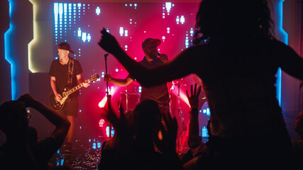 Rock Band Performing a Song at a Concert in a Night Club. Front Row Crowd is Partying. Fan Girl is Sitting on Shoulders of Her Boyfriend in Front of Bright Colorful Strobing Lights on Stage. 