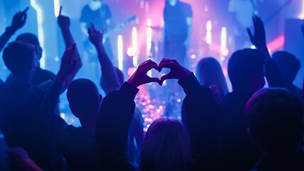 Fototapeta na wymiar Person is Making a Heart Sign Gesture and Holding Hands Up at a Performance. Rock Band Playing a Song at a Concert in a Night Club on Stage with Bright Colorful Strobing Lights.