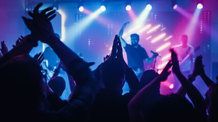Fototapeta na wymiar Rock Band with Guitarists and Drummer Performing at a Concert in a Night Club. Front Row Crowd is Partying. Silhouettes of Fans Clap Hands in Front of Bright Colorful Strobing Lights on Stage. 