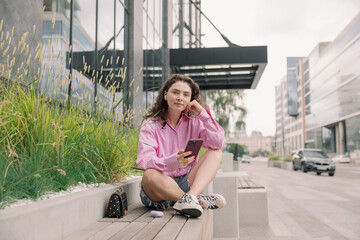 Portrait of young woman listening to music with mobile phone and relaxing while sitting on bus stop.