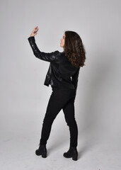 Full length portrait of young woman with natural brown hair,  wearing black leather scifi outfit with jacket, standing pose  with back to the camera, on light studio background.