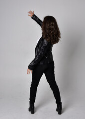 Full length portrait of young woman with natural brown hair,  wearing black leather scifi outfit with jacket, standing pose  with back to the camera, on light studio background.