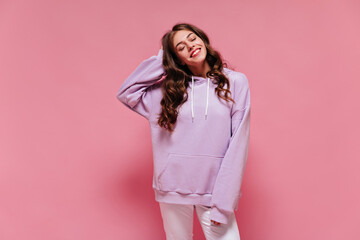 Brunette happy woman in purple sport suit smiles sincerely. Curly young girl in hoodie and light pants touches hair on pink background.