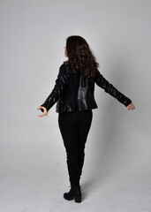 Full length portrait of young woman with natural brown hair,  wearing black leather scifi outfit with jacket, standing pose  with back to the camera, on light studio background.