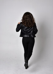 Full length portrait of young woman with natural brown hair,  wearing black leather scifi outfit with jacket, standing pose  with back to the camera, on light studio background.