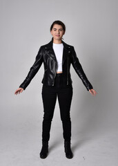 Full length portrait of young woman with natural brown hair,  wearing black leather scifi outfit with jacket, standing pose on light studio background.