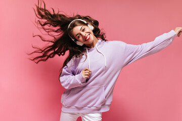 Pretty woman in oversized purple hoodie dances on isolated pink background. Brunette girl listening...