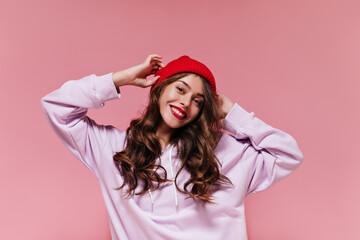Charming cool girl in purple hoodie puts on red hat. Pretty brunette curly woman smiles widely on...