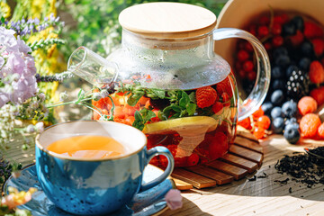 natural aromatic fruit tea in a transparent teapot and in a cup among berries and flowers. warming aromatic tea with a deep aroma of berries and wildflowers.