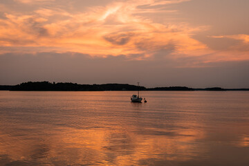 Naklejka na ściany i meble Beautiful sunset on the Baltic Sea. Amazing golden orange pink tones of the sunset cloudy sky. Islands of archipelago reflections on water surface. Small fishing boat is anchored off the coast.