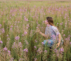 Beautiful smiling blond young woman in light purple shirt sitting on field of fireweed flowers sniffing the scent of pink flower copy space enjoing the nature