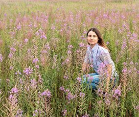 Young beautiful blond woman in purple shirt sitting in the meadow among flowers of fireweed, beauty in nature landscape enjoing the nature