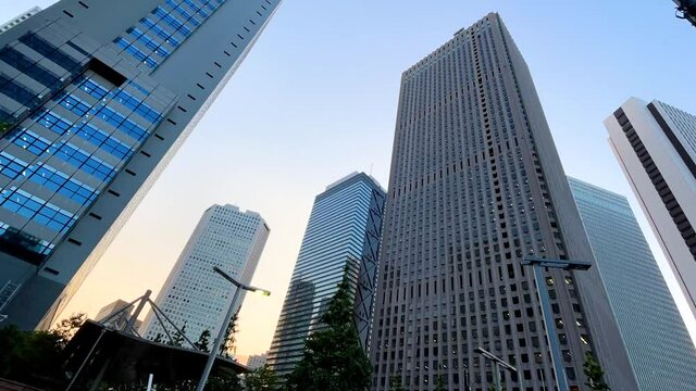SHINJUKU, TOKYO, JAPAN - JUNE 2021 : Exterior of tall office buildings in sunrise time. Wide low angle time lapse zoom in shot, morning to day. Japanese urban metropolis and business concept shot.