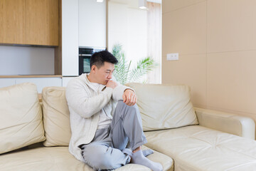 Bored friends young asian man sitting on sofa at home alone. Misses male sitting on the couch in apartment. Lonely guy indoors living room. Worried and stressed about problems in the family or at work