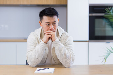 Naklejka premium Thoughtful serious young asian man looking away. student writer sit at home office desk with laptop thinking of inspiration search solution ideas lost in thoughts dreaming solving financial problems