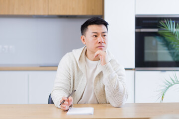 Thoughtful serious young asian man looking away. student writer sit at home office desk with laptop thinking of inspiration search solution ideas lost in thoughts dreaming solving financial problems