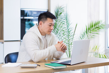 Young asian man working at home on a laptop computer sitting at a table in the kitchen