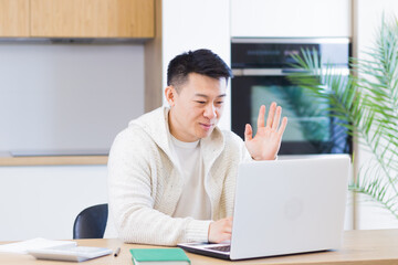 young asian man sitting at home in the living room or kitchen and communicating online. Male waving hand at laptop webcam. Video call learning distance. Training, conference or meeting E-learning