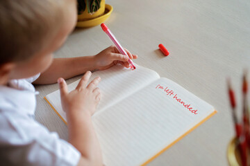 Happy left-handed boy writing in the paper book with his left hand, international left-hander day...