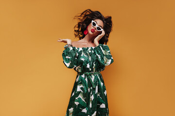 Unusual curly brunette haired girl with white sunglasses and red earrings in fashionable cool outfit blowing kisses and looking into camera..