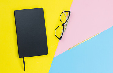 Black notebook, pen, glasses on yellow, pink and blue paper background. Designer office desk table....