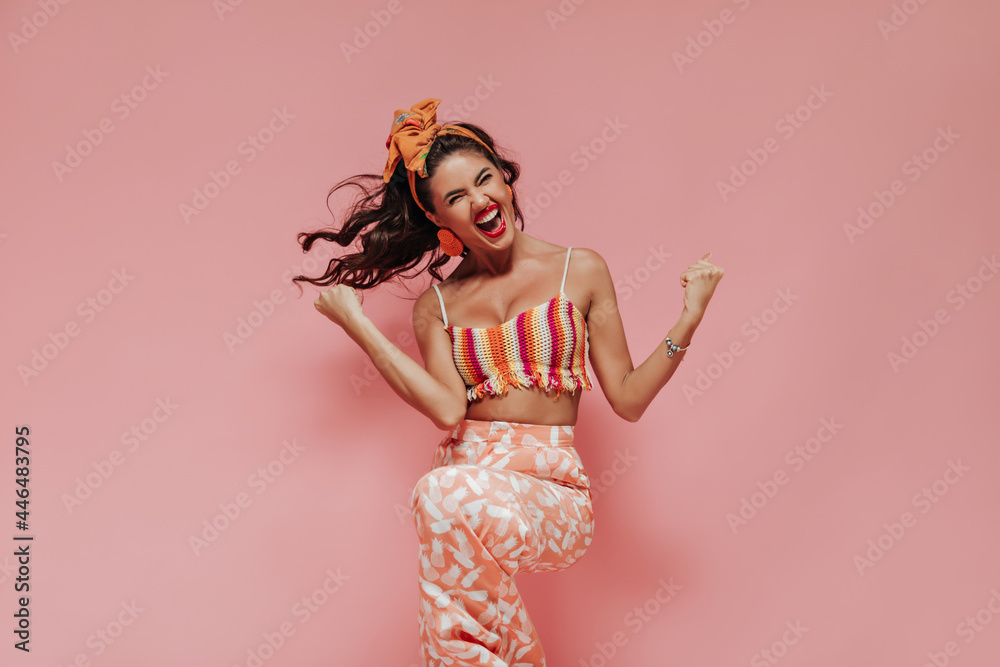 Wall mural Emotional curly haired woman with red lipstick in striped bright top and pink cool trousers rejoices on isolated background..