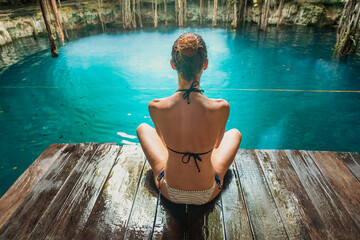 Mujer sentada en plataforma de madera mirando el agua de un cenote en Mexico - obrazy, fototapety, plakaty