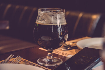 Luxury still life with elegant glass of stout beer and smartphone atop table in restaurant. Low key glassware closeup