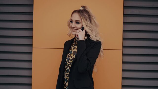 The sexy, blonde young woman, with bright make-up, talks on the phone, displays a wide smile, isolated on an orange and brown background. Female energy. Beauty industry. Artistic concept