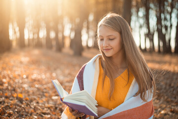 Young blond girl reading a book sitting on the bench in autumn park with bright sun on the background. Bright warm photo of a girl with a book in autumn park covered with yellow foliage