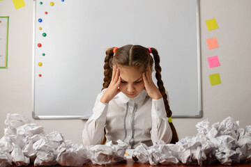Concept of brainstorming, thinking hard, failure, difficult decision, doing difficult task. Little girl in the classroom sitting around many crumpled papers - Powered by Adobe