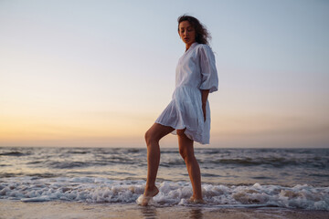 Young happy woman in a white fluttering dress walks along seashore. The girl looks at the magical sunrise. Summer time. Travel, weekend, relax and lifestyle concept.