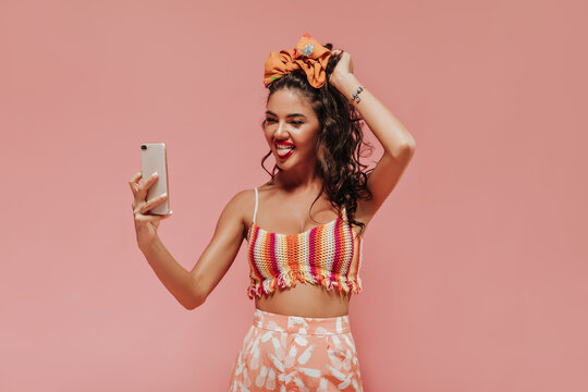 Funny curly girl with stylish headband and accessories in colorful top and pineapple print pants makes photo on pink backdrop..