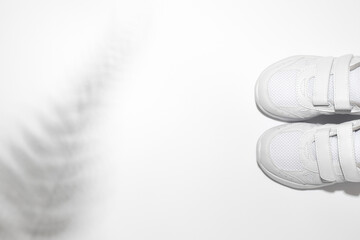 flat lay two white child sneakers with velcro fasteners with shadows from a fern or palm tree isolated on a white background.