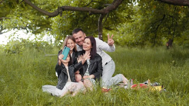 Cheerful caucasian family of four taking selfie on modern smartphone during picnic time. Young parents with two cute daughters taking fun. Parents with kids using mobile for taking selfie