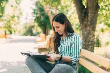 Fototapeta premium Photo of smiling young woman sitting on bench with pen and book, in park and writing in planner