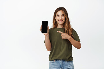 Portrait of smiling adult woman pointing finger at screen of mobile phone, showing interface, recommend application, website of store or company, new feature in app, white background