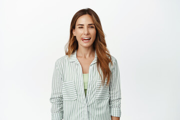 Cheerful brunette woman winking and showing tongue, having fun, looking happy, standing in blouse against white background