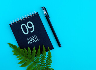 April 9th. Day 9 of month, Calendar date. Black notepad sheet, pen, fern twig, on a blue...