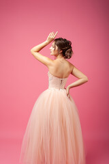 Fototapeta na wymiar A young woman stands with her back against an isolated background. A beautiful woman model in a beige ball gown and evening hairstyle with bent arms poses on a pink background.