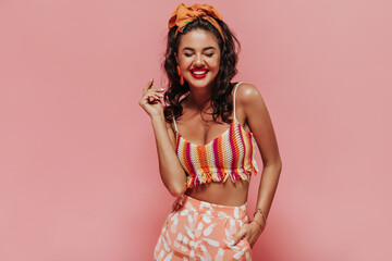 Smiling trendy woman with orange headband and red lipstick in striped modern top and printed pants...