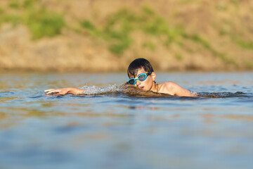 Happy boy in swimming glasses swims in the river in the summer at sunset. A child enjoys a summer children's holiday on the shore of the lake. Active holidays. Dynamic Image