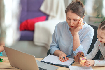 tired young woman works at home using laptop, together with her little daughter. A naughty child prevents his mother from working and wants to communicate with his mother, make noise and play nearby.
