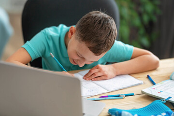 inquisitive boy uses laptop to study school subjects during his online lesson at home. Writing down topic lesson in notebook. E-Education Distance Home Education.