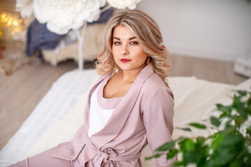 Naklejka premium Portrait of an attractive beautiful young woman in an elegant outfit in a bright interior. a pretty girl with curly blonde hair, in a beautiful dress, with beautiful makeup and styling looks away.
