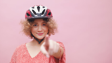 Beautiful young girl wearing a bicycle helmet smiling and pointing at the camera. The girl is dressed in a casual pink top. Cycling for leisure healthy lifestyle concept. Isolated over pink background - obrazy, fototapety, plakaty