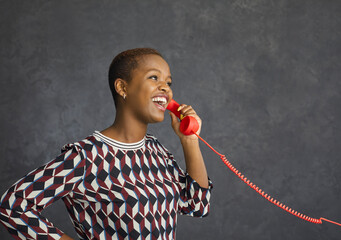 Studio portrait of happy young black woman talking on red fixed land line telephone on grey...