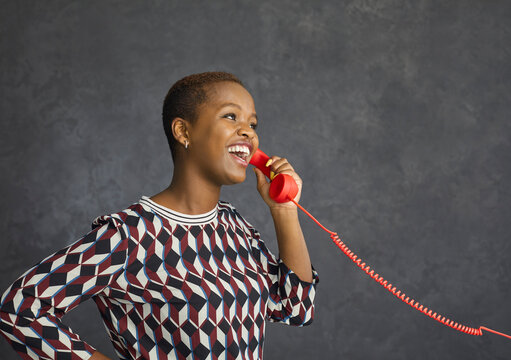 Studio Portrait Of Happy Young Black Woman Talking On Red Fixed Land Line Telephone On Grey Background. Positive Cheerful Carefree Customer Answers Phone Or Calls Hotline Client Support Service Center